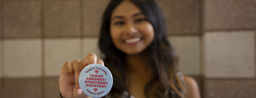 Image of student holding up a button pin that says "Rutgers University Student Affairs - Proud Resident/Apartment Assistant"